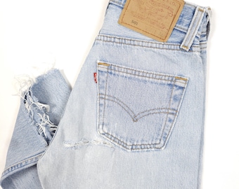 Puede incluir: Vaqueros de mezclilla azul claro con detalles desgastados, incluyendo una sección rasgada en la pierna. Los vaqueros presentan un diseño clásico de cinco bolsillos, una etiqueta roja de Levi's y un parche de cuero marrón con el logo de la marca.