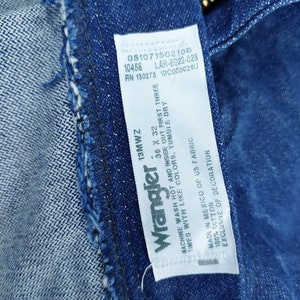 May include: A white Wrangler jeans care label with blue text. The label includes the size 38 x 32, the fabric content, and washing instructions. The label also includes the following numbers: 05107150210в, 10458, LAR-E022-023, RN 130273, 100000260, and 13MWZ.