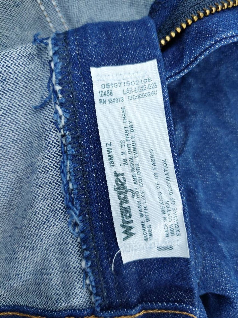 May include: A white Wrangler jeans care label with blue text. The label includes the size 38 x 32, the fabric content, and washing instructions. The label also includes the following numbers: 05107150210в, 10458, LAR-E022-023, RN 130273, 100000260, and 13MWZ.