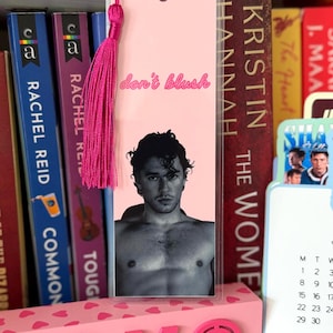 May include: A rectangular bookmark with a light pink background featuring the text "don't blush" in a pink script font. Below the text is a black and white photograph of a shirtless person. A bright pink tassel is attached to the top hole.