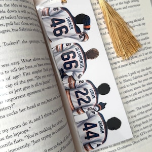 May include: A rectangular white bookmark featuring four illustrated hockey players wearing jerseys with the names Tucker 46, Di Laurentis 66, Logan 22, and Graham 44. The bookmark is topped with a gold-colored decorative tassel and is placed inside an open book.