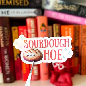 May include: A sticker with the text "SOURDOUGH HOE" in red letters, featuring a cartoon loaf of bread with a smiling face. The sticker has a white border and is held by tweezers in front of a bookshelf.