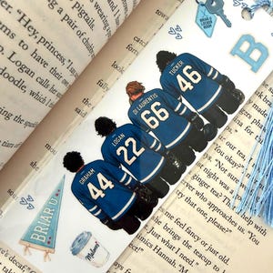 Könnte beinhalten: Ein rechteckiges Lesezeichen mit vier Eishockeyspielern in blauen Trikots mit den Namen und Nummern Graham 44, Logan 22, Di Laurentis 66 und Tucker 46. Es enthält einen Briar U Wimpel, einen Malones Kaffeebecher, einen Schlüsselanhänger und eine hellblaue Quaste.