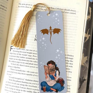 May include: A rectangular bookmark with a light blue background featuring a gold dragon silhouette, white star accents, and an illustration of a person reading a book titled Fourth Wing, finished with a gold tassel attached at the top.