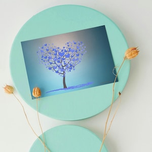 Puede incluir: Una impresión rectangular que muestra un árbol con hojas azules en forma de corazón sobre un fondo azul degradado, colocada sobre una superficie redonda de color verde menta con tallos botánicos secos en primer plano.