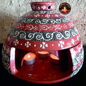 May include: Red terracotta candle holder with black and white painted designs. The holder has three openings for tealight candles. The text "CANDLE LIGHT HANDMADE TERRACOTTA ART" is on the top of the holder.