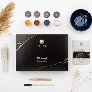 Pode incluir: Um kit Kintsugi Deluxe Box da Chiyu Fine Arts & Crafts contendo uma caixa preta com linhas douradas, quatro pequenos frascos de pó colorido, uma tigela de cerâmica azul com linhas de reparação douradas, um manual, um tubo de cola e dois pincéis.