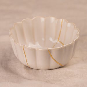 May include: A white ceramic bowl with a scalloped rim and vertical ridges. The bowl features gold-colored lines that highlight the repaired cracks, showcasing the Japanese art of Kintsugi. The bowl is a decorative piece.