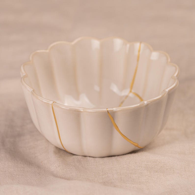 May include: A white ceramic bowl with a scalloped rim and vertical ridges. The bowl features gold-colored lines that highlight the repaired cracks, showcasing the Japanese art of Kintsugi. The bowl is a decorative piece.
