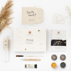 May include: A Kintsugi Modern Repair Kit in a flat lay. The kit includes a box with the text "Kintsugi Modern Repair Kit Gold & Silver", a small bag with "Perfectly Imperfect" text, and various repair materials like epoxy putty and brushes.