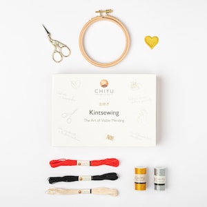 Può includere: Un kit artigianale per rammendi visibili, con un telaio da ricamo in legno, forbici dorate e una toppa dorata a forma di cuore. Il kit include una scatola con il testo "Kintsewing The Art of Visible Mending", insieme a rocchetti di filo rosso, nero e crema, e filo dorato e argentato.
