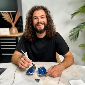 May include: A person smiling while working on a broken blue ceramic bowl. The bowl is in pieces on a marble table, with a paintbrush in hand. The bowl has a dark blue glaze with a white rim.