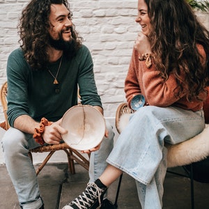 May include: A handmade ceramic bowl with a visible crack, held by a person. The bowl is off-white with a brown crack. The person is wearing a green long-sleeve shirt and light blue jeans. Another person is wearing an orange sweater and light blue jeans.