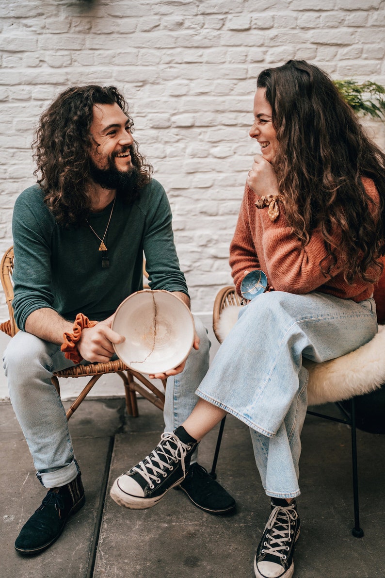 May include: A handmade ceramic bowl with a visible crack, held by a person. The bowl is off-white with a brown crack. The person is wearing a green long-sleeve shirt and light blue jeans. Another person is wearing an orange sweater and light blue jeans.