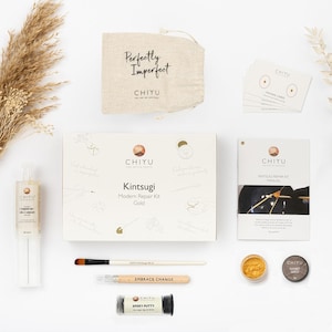 May include: A flat lay of a Kintsugi Modern Repair Kit in gold. The kit includes a box, a booklet, a brush, epoxy putty, mixing cards, and a small bag with the text "Perfectly Imperfect". The kit is designed for repairing broken items.
