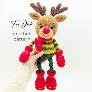 Op de afbeelding: Gehaakte rendierpop met een rode neus, geweien en een groen-gele gestreepte trui. Het rendier draagt rode wanten, een rode sjaal en rode laarzen. De tekst "For Didi crochet pattern" is zichtbaar.