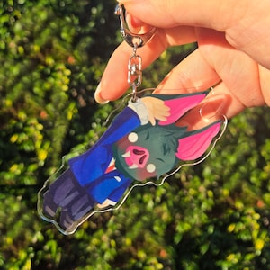 May include: A clear acrylic keyring featuring a cartoon bat character with dark fur, large bright pink ears, and white fangs. The character is wearing a blue suit jacket and dark trousers, depicted in a dynamic pose with one arm raised.