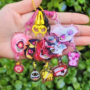 May include: A set of eight colourful character charms on keychains. The charms feature cartoon characters with pink, red, white, black and yellow accents. The keychains are attached to a silver chain.