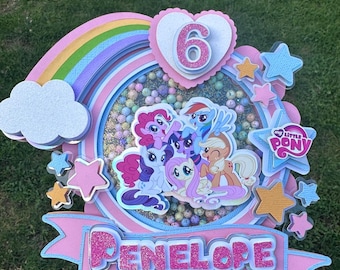 May include: A pink and blue My Little Pony cake topper with a rainbow, a cloud, stars, and the name Penelope. The topper features a heart with the number 6 inside.