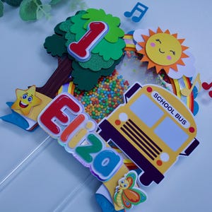 Könnte beinhalten: Ein bunter Tortendekor mit einem Schulbus, einem Regenbogen, einer Sonne, einem Baum, einem Stern, Noten und dem Wort "First".