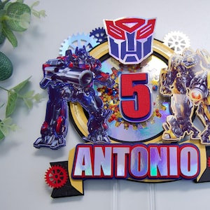 Pode incluir: Um topo de bolo com tema Transformers com dois robôs, um número 5 vermelho e o nome "ANTONIO" em letras iridescentes. Os robôs são azuis e amarelos com detalhes prateados. O topo é decorado com engrenagens e outros detalhes mecânicos.