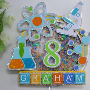 May include: A colourful science-themed cake topper featuring a beaker, atom, microscope, and molecule. The number 8 is in the centre, and the name "GRAHAM" is spelled out using periodic table elements.