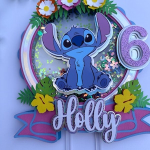 Könnte beinhalten: Ein blauer Stitch-Charakter-Kuchentopper mit einer glitzernden 6 in Lila, umgeben von Blumen und Blättern. Der Topper enthält auch ein rosa Banner mit dem Namen "Holly" in glitzernden lila Buchstaben.