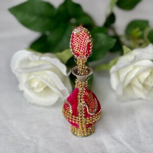 May include: A small, ornate perfume bottle with a pink velvet body and gold accents. The bottle is decorated with sequins and beads. The bottle is sitting on a white fabric surface.