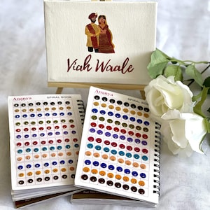 May include: Two spiral bound books with colour swatches of different shades of fabric. The books are titled "Ananya Spiral Book" and are on a white surface with a white canvas sign that says "Viah Waale" with a drawing of a bride and groom in traditional Indian clothing.