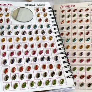 May include: A spiral-bound book featuring a variety of Kumkum Bindi designs. Each Bindi is oval-shaped with a central color and a gold-colored border. The colors include red, yellow, blue, green, and pink. A small mirror is also visible.