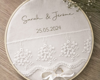 May include: White fabric ring pillow with lace trim and embroidered flowers. The pillow has the text "Sarah & Jerome" and "25.05.2024" embroidered on it. Two gold rings are tied with a white satin ribbon.
