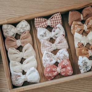 Może przedstawiać: Drewniane pudełko wypełnione 12 ręcznie robionymi kokardkami do włosów z tkaniny. Kokardki są w różnych wzorach i kolorach, w tym kwiatowych, w kratkę i jednokolorowych. Kokardki są ułożone w rzędach i gotowe do wysyłki.