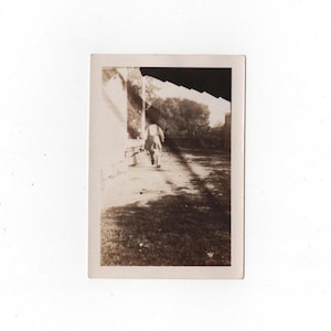 May include: A vintage sepia-toned photograph showing a person walking away from the camera along a path beside a building. The scene is captured in natural light with long shadows cast across the ground, featuring trees in the background.