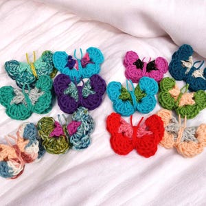May include: A collection of colorful, crocheted butterfly ornaments in various shades of blue, green, purple, pink, and red. The butterflies are arranged on a white surface, showcasing their intricate handmade details. Each butterfly has a unique color combination.