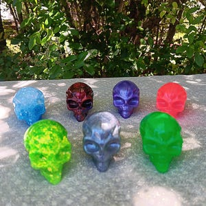 May include: A collection of seven alien skull figurines in various colors, including blue, red, purple, green, and silver. The skulls are arranged on a gray surface with a natural green background.