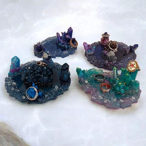 May include: Four decorative ring holders with crystal and gemstone accents. Each holder features a unique color palette, including shades of blue, purple, and green. Rings of various styles are displayed on the holders, showcasing their design.