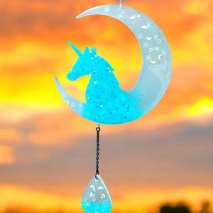 May include: A blue unicorn silhouette inside a crescent moon with a teardrop-shaped pendant hanging from the bottom. Both the moon and pendant are filled with glitter.