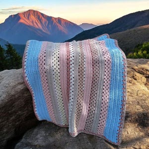 May include: A handmade crochet blanket in pastel colors of blue, pink, and cream, draped over a rock. The blanket features vertical stripes and a textured stitch pattern. The background shows a mountain range at sunset.