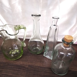 May include: Four glass bottles of varying shapes and sizes. The bottles are clear and have a green tint. The bottles are sitting on a wooden surface.