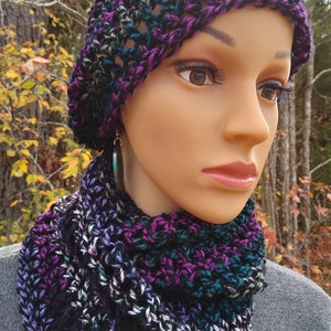 May include: A handmade crochet hat and cowl set in shades of purple, teal, and black. The open weave design of the hat and cowl creates a textured look. The cowl is wrapped around a mannequin's neck, showcasing the color variations.