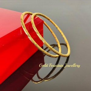 May include: Two gold bangles with a textured design. The bangles are stacked on top of each other and are resting on a red box.