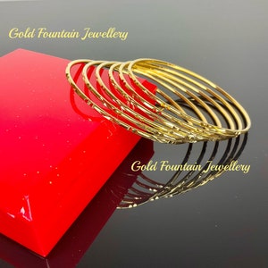 May include: A set of seven gold bangles with a textured surface. The bangles are stacked on top of each other and are on a red surface. The text "Gold Fountain Jewellery" is visible in the image.