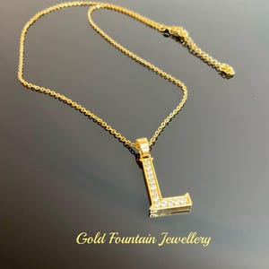 May include: A gold chain necklace with a pendant in the shape of the letter L. The pendant is encrusted with clear cubic zirconia stones. The necklace is on a black background. Gold Fountain Jewellery.
