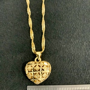 May include: A gold heart-shaped pendant necklace with a detailed, intricate design. The pendant is approximately 2.5 cm in diameter. The chain is a twisted rope style.