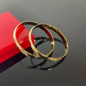 May include: Two gold-toned metal bangle bracelets with a polished finish and a textured, glittery section on the outer surface. The bangles are circular, thin, and feature a hinged clasp mechanism visible on the side of the metal bands.