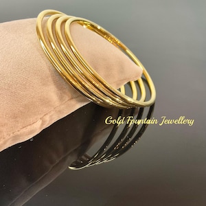 May include: Five gold bangle bracelets stacked on top of each other on a light brown velvet surface. The bracelets are all the same width and have a smooth, polished finish. The text "Gold Fountain Jewellery" is visible at the bottom of the image.