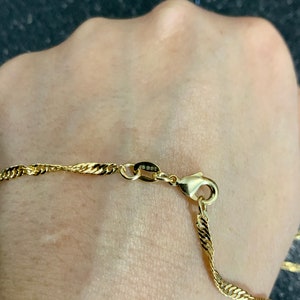 May include: A gold chain necklace with a twisted rope design. The chain has a lobster clasp closure. The chain is on a person's wrist.
