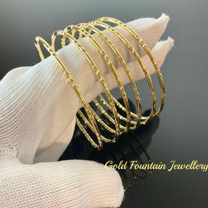 May include: A set of six gold bangles with a textured design. The bangles are stacked on top of each other and are displayed on a white surface. Gold Fountain Jewellery is printed on the surface.