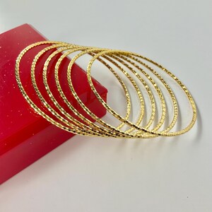 May include: A set of six thin, circular gold bangles featuring a textured, faceted surface that reflects light. The bangles are arranged in a stacked, overlapping formation, creating a series of concentric rings with a bright, metallic finish.