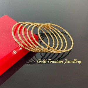 May include: A set of six gold-toned bangles with a textured design. The bangles are stacked on top of each other and are arranged in a semi-circle. The text "Gold Fountain Jewellery" is visible in the bottom right corner of the image.